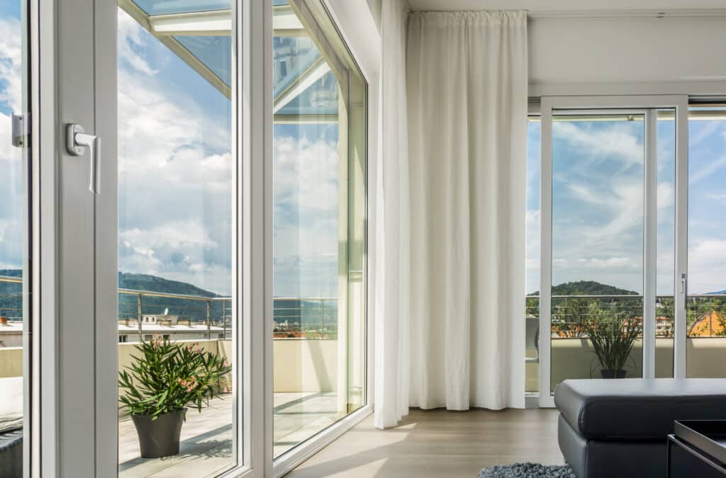 Intérieur moderne avec grandes baies vitrées donnant sur une terrasse ensoleillée et vue sur les montagnes. Rideaux blancs et canapé en cuir noir.