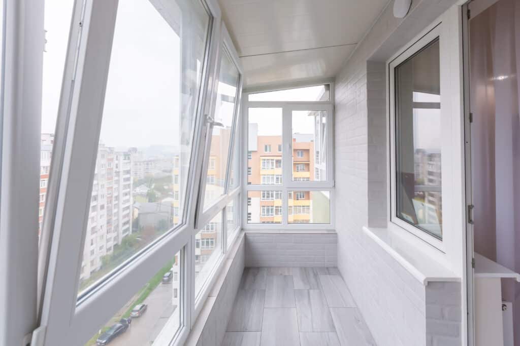 Balcon moderne vitré avec grandes fenêtres blanches ouvertes, sol carrelé gris clair et vue dégagée sur les immeubles urbains.