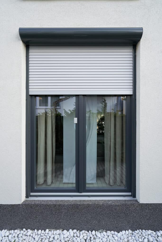 Fenêtre moderne sur façade blanche avec volet roulant gris à moitié fermé. Des galets blancs décorent le pied du mur.