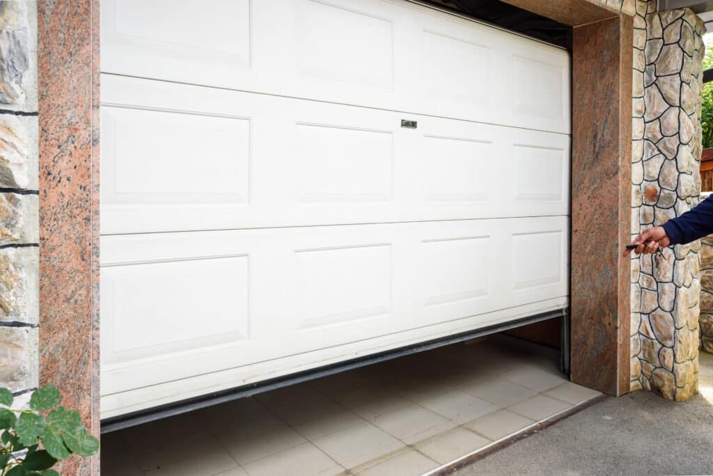 Main ouvrant une porte de garage blanche automatique avec une télécommande. Le sol carrelé du garage est visible. Piliers en pierre et granite.