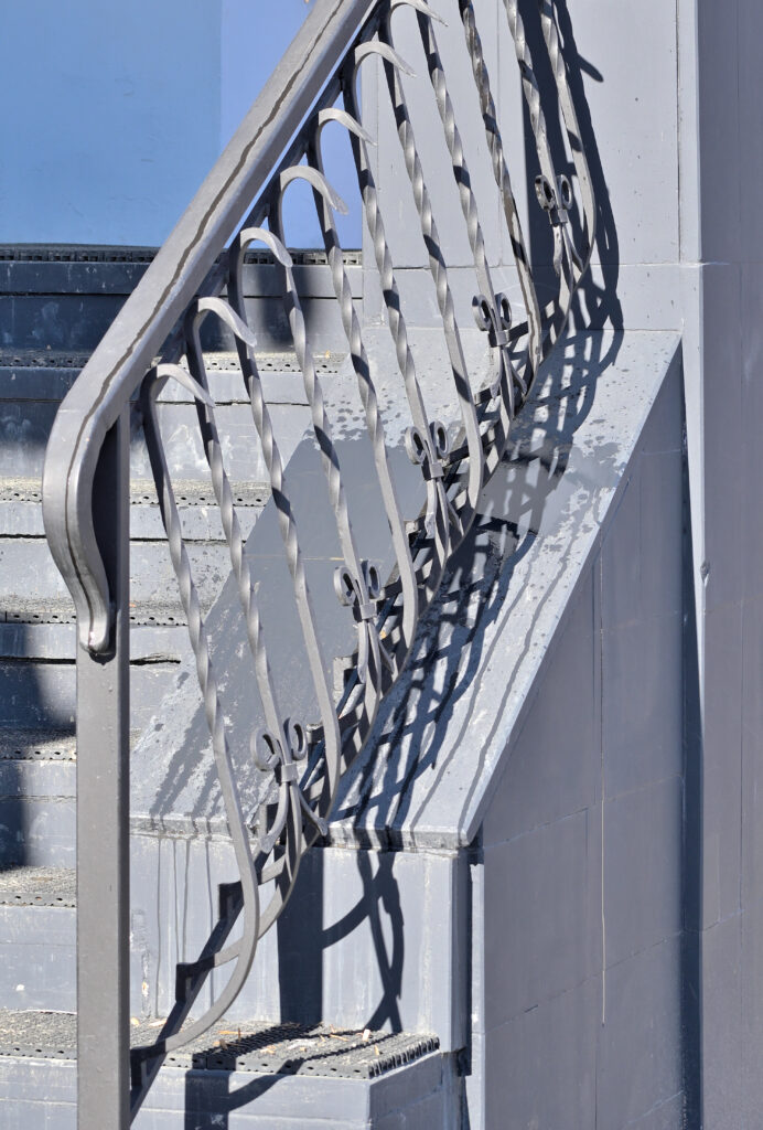 Rampe d'escalier en métal gris foncé avec motifs forgés, projetant des ombres graphiques sur les marches et le mur bleu clair.