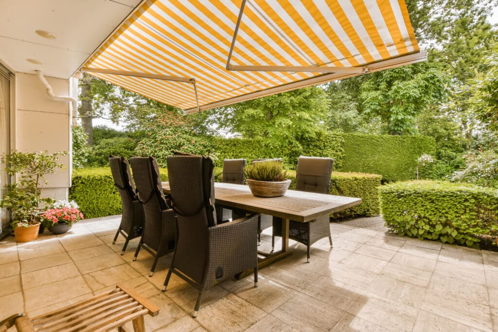Grande terrasse carrelée avec table à manger en bois, chaises en osier et auvent jaune et blanc rayé, entourée d'un jardin luxuriant.
