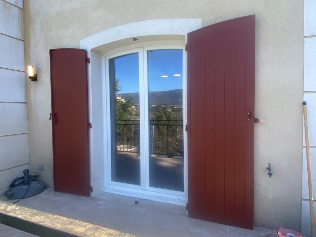 Volets rouge-brun ouverts sur une porte-fenêtre blanche. Terrasse avec vue panoramique sur une vallée montagneuse ensoleillée.